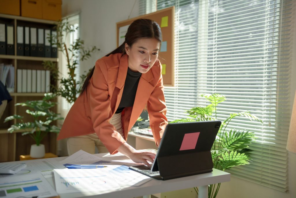 Young professional woman wearing an orange jacket leaning on her desk while using a tablet and analyzing financial charts in her office, sunlight coming through the window blinds